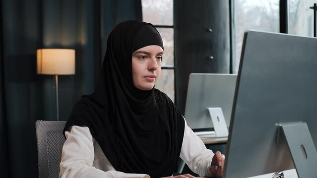 Muslim focused female executive manager businesswoman sitting at desk working typing message on laptop pc in contemporary corporation office. Data Analysis, plan strategy, business technologies.
