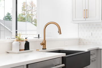 Fototapeta na wymiar A beautiful sink in a remodeled modern farmhouse kitchen with a gold faucet, black apron or farmhouse sink, white granite, and a tiled backsplash. 