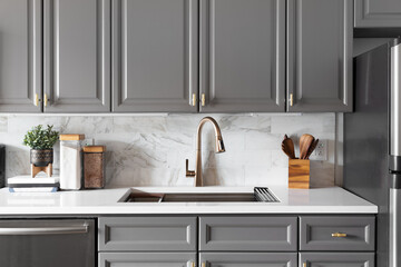 A kitchen sink detail shot with grey cabinets, a white marble countertop and backsplash, and...