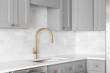 A kitchen sink detail shot with a gold faucet, marble backsplash, grey cabinets, and gold hardware.