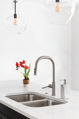 A kitchen detail shot with glass pendant lights hanging above a kitchen island with a stainless steel faucet and sink, marble countertop, and red flowers.
