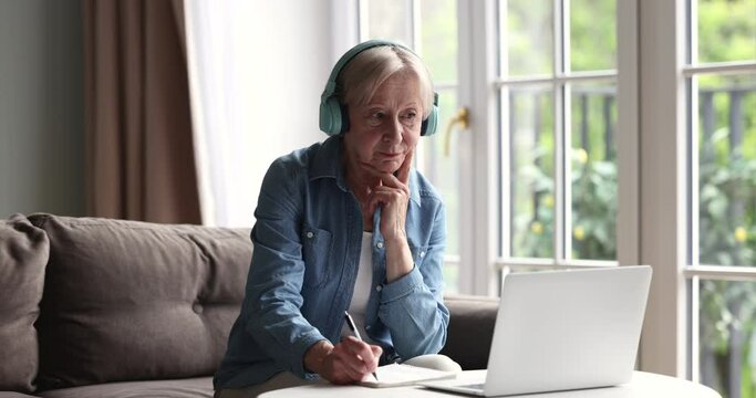 Attractive senior woman in headphones jotting information, makes notes in copybook, listen online teacher, get new knowledge and skills on retirement use laptop. Older generation and modern tech usage