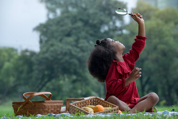 Happy little girl in a red dress, using magnifying glass and looking up on the green lawn