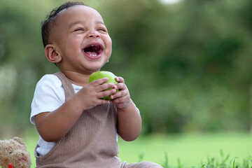 Adorable funny African baby boy enjoy with green apple on blanket in a green summer garden on a sunny morning
