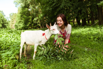 Woman dressed traditional fashionable ukrainian embroidery vyshyvanka dress shirt ethnic costume cloth feeding goat with leaves grass in the forest village. Ukrainian culture