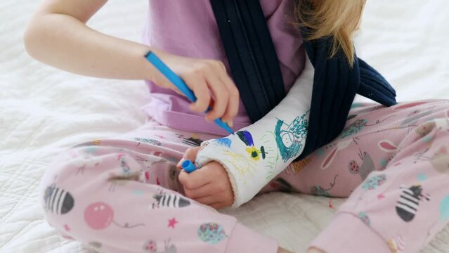 Adorable preschooler girl with a broken arm at home on the bed draws with felt-tip pens on an orthopedic cast. How to have fun with a broken limb. Broken arm in a cast in kids