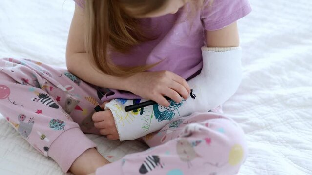 Adorable preschooler girl with a broken arm at home on the bed draws with felt-tip pens on an orthopedic cast. How to have fun with a broken limb. Broken arm in a cast in kids