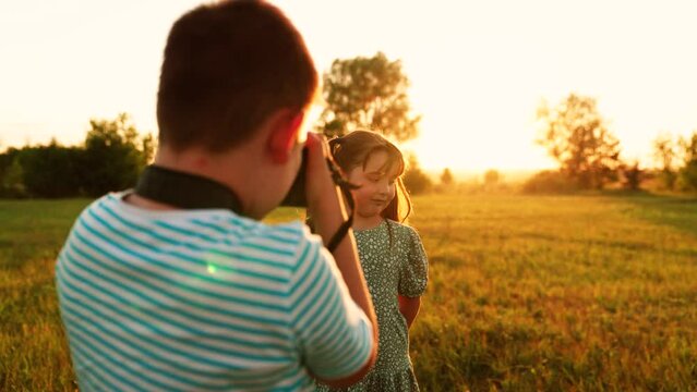 Child girl poses as model for boy to photographer with camera. Children are photographed in park at sunset with digital camera. Childrens games in nature in sun. Hobby of children is taking pictures
