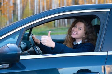 Portrait of happy positive girl, young woman driver is sitting in her car, new automobile, enjoying driving, having fun, laugh. Joyful lady in auto showing thumb up, like gesture