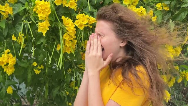 Portrait of beautiful young allergic woman is suffering from pollen allergy or cold on natural flower flowering tree background at spring or sunny summer day sneezes, blowing her runny nose rubs eyes