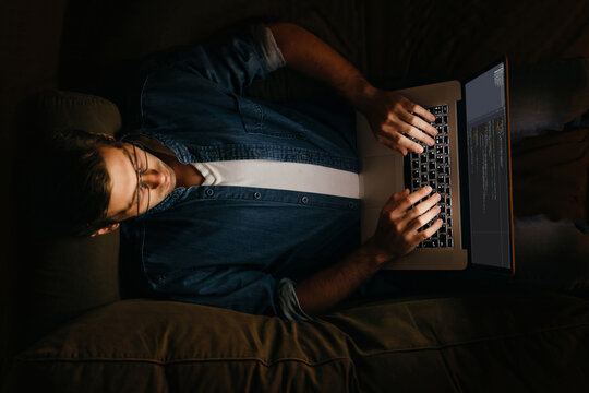 Top view of male programmer laying on sofa and working at his laptop. Overworked coder working from home at night. 