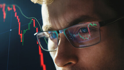 Close-up portrait of focused crypto trader analyst wearing eyeglasses looking at computer screen...
