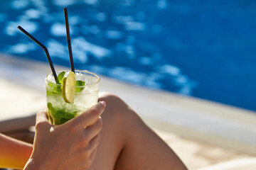 Glass of cool cocktail in glass with ice in hand by pool against background blue sparkling water. Girl with cocktail in hand is sunbathing sunny day by pool on territory of hotel in resort on vacation