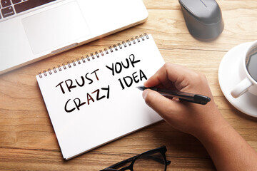 Trust Your Crazy Idea, motivational words and sentences for work and life. Quote sentence in notebook with laptop, pen, coffee over wooden background.