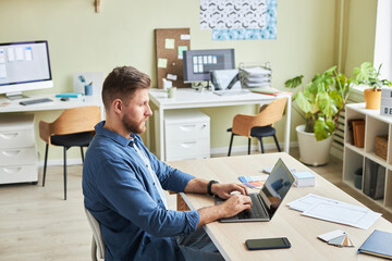 Side view portrait of bearded young man using laptop while working in cozy office, copy space