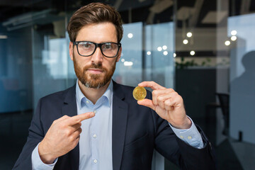 Serious and focused businessman looking at camera, mature successful investor holding bitcoin coin cryptocurrency and pointing, bearded man working inside modern office.