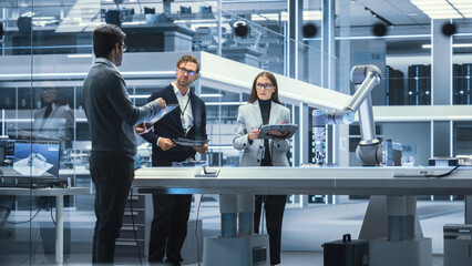 Diverse Team of Industrial Robotics Engineers Gathered Around a Table With Robotic Arm. Scientists Use Tablet Computer to Manipulate and Program the Robot to Pick Up and Move a Metal Component.