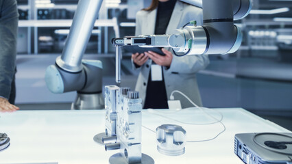 Close Up Shot of a Modern Robotic Arm Holding and Moving a Mechanical Metal Component. Diverse Team of Engineers Using Tablet Computer to Manipulate and Program the Robot.