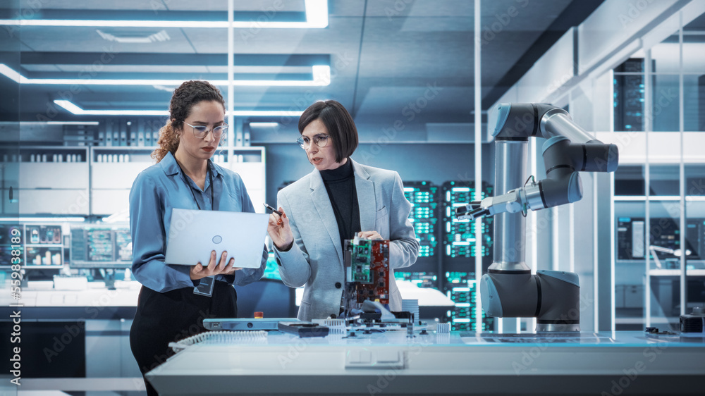 Poster Two Female Working in High Tech Facility. Robotics Engineer and Machinery Operator Collaborate on a Robot Arm Project, Using a Laptop Computer to Operate the Robot Arm in Technology Startup Lab.