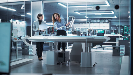 Two Female Working in High Tech Facility. Robotics Engineer and Machinery Operator Collaborate on a Robot Arm Project, Using a Laptop Computer to Operate the Robot Arm in Technology Startup Lab.