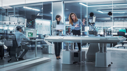 Professional Multicultural Team of Two Female Engineers Using Industrial Programmable Robotic Arm in a Factory Development Workshop. Robot Hand Machine Picking Up and Holding a Microchip.