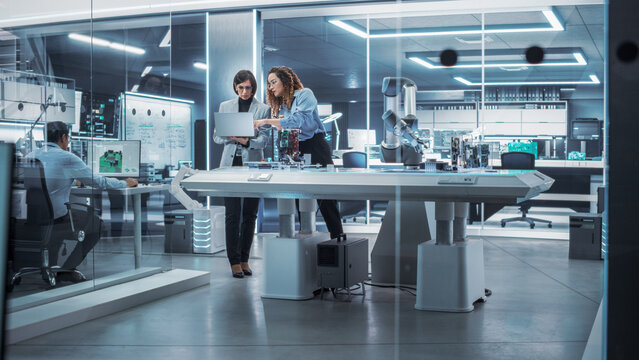 Young Female Engineer Works with Colleague Robotics Scientist with Laptop Computer. They Collaborate on a Robot Arm Development in an Industrial Startup. Analyzing and Evaluating Production Capacity.