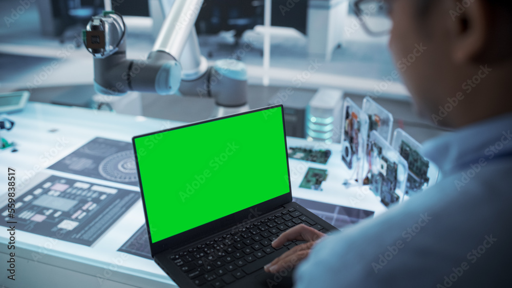 Fototapete Over the Shoulder Shot of a Robotics Engineer Using a Laptop Computer with Green Screen Mock Up Display Screen. Robotic Arm Holding a Microchip. Automation Startup Research and Development Office.