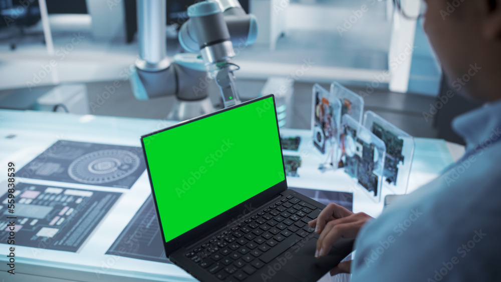 Fototapete Over the Shoulder Shot of a Robotics Engineer Using a Laptop Computer with Green Screen Mock Up Display Screen. Robotic Arm Holding a Microchip. Automation Startup Research and Development Office.