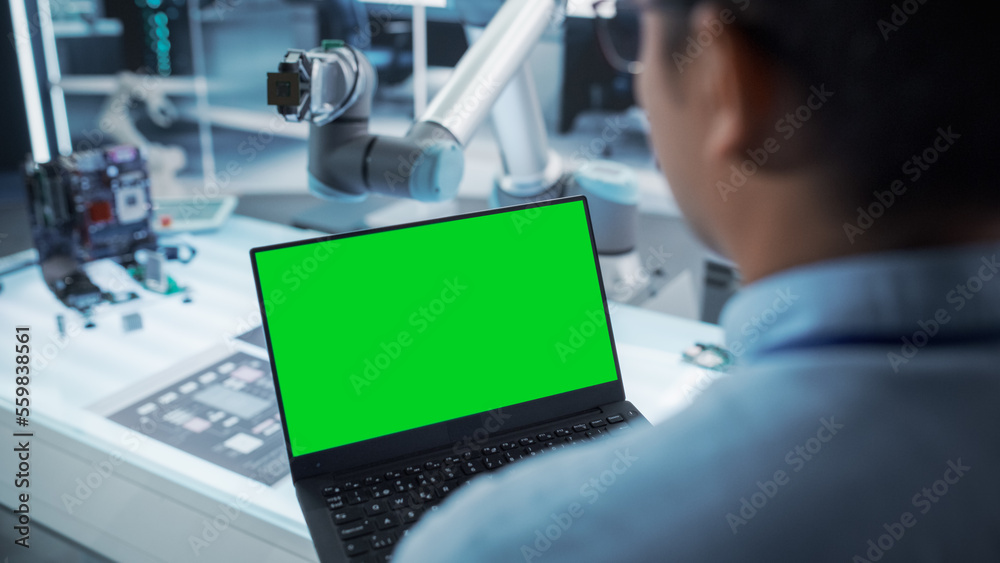 Aufkleber Over the Shoulder Shot of a Robotics Engineer Using a Laptop Computer with Green Screen Mock Up Display Screen. Robotic Arm Holding a Microchip. Automation Startup Research and Development Office.