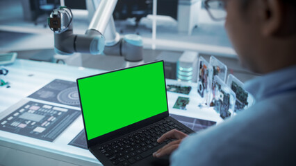 Obraz na płótnie Canvas Over the Shoulder Shot of a Robotics Engineer Using a Laptop Computer with Green Screen Mock Up Display Screen. Robotic Arm Holding a Microchip. Automation Startup Research and Development Office.