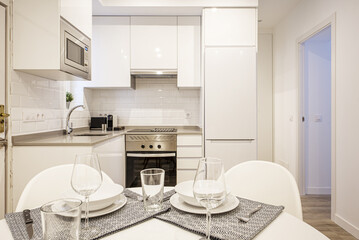 Living room with a round dining table with tableware set with crystal stemware and white crockery in an open plan kitchen with white gloss cabinets and gray stone worktops