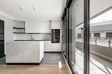 Fototapeta na wymiar Newly installed modern kitchen with central island, smooth gray cabinets, black glass appliances and glass-covered wall with access to an open terrace