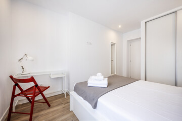 Bedroom with white wooden bed, gray blanket, clean white towels, built-in wardrobe with sliding doors and desk with metal and glass top and wooden folding red chair