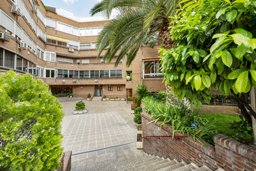 Facade of a four-story building with white aluminum windows and walkways with grass, slats and a recreation area with flowers and tree-lined gardens