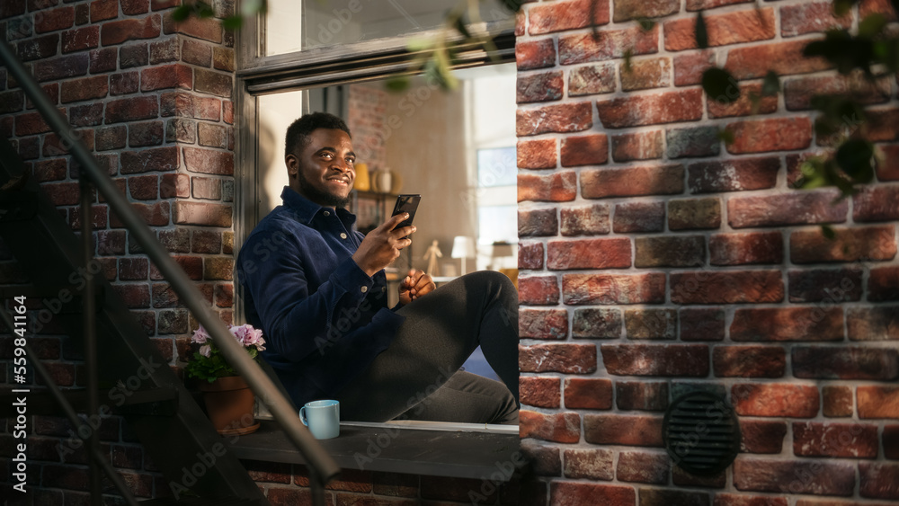 Aufkleber black authentic man using smartphone in his flat while sitting on bedroom windowsill. african americ