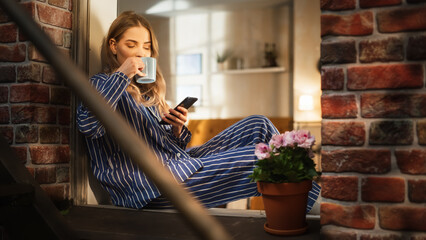 Young Beautiful Woman Using Smartphone and Enjoying her Morning Coffee While Sitting on her Bedroom...