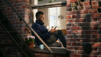 Black Handsome Man Using his Smartphone While Enjoying the View from his Bedroom Window. Young Male Checking his Social Media While Drinking his Coffee and Relaxing in Brooklyn Style Brownstone House