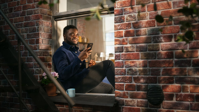 Black Authentic Man Using Smartphone in his Flat While Sitting on Bedroom Windowsill. African American Male Posting Photos on Social Media and Using Filters While Smiling. Calm and Relaxing Evening.