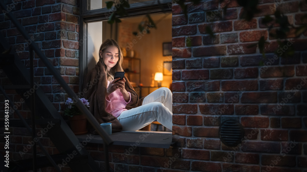 Poster happy beautiful white woman using her smartphone while sitting on her windowsill during evening. she