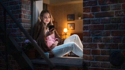 A Beautiful White Woman is Using her Smartphone While Sitting on her Windowsill at Night With a...