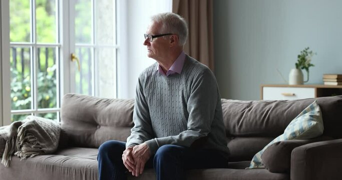 Sad unhappy elderly retired man sits on couch alone, sighs, looks out window feels abandoned, suffer from weakness, ageing disorders, grieving about past, missing for grown up children living far away