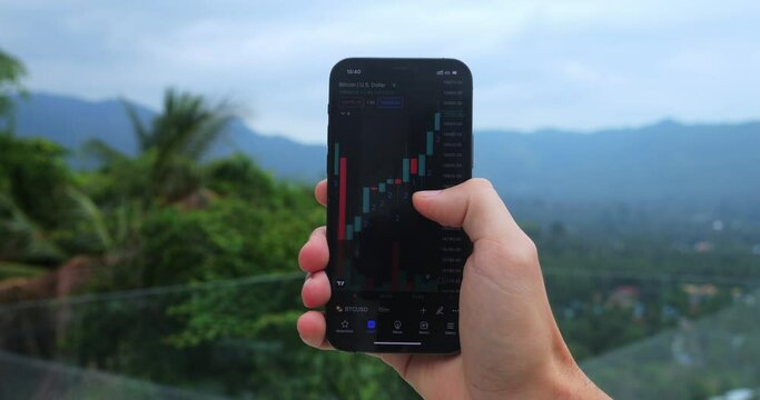 Close-up of male crypto enthusiast holding smartphone app studying graph of changes in the value of cryptocurrency on the stock exchange on backdrop of beautiful green nature, mountains and palms.
