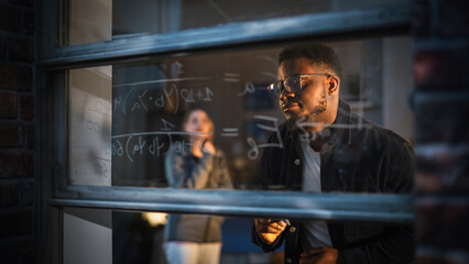 African American Young Man Using an Erasable Marker to Solve a Math Equation on Window Glass. Black Male Tutor Writing a Physics Theory and Explaining it to Fellow Female Student.