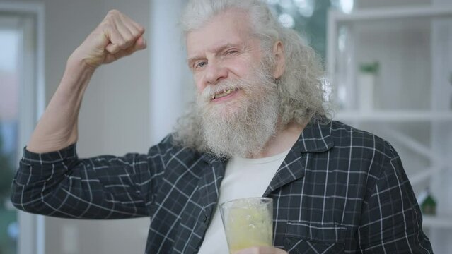 Handsome Senior Caucasian Man Drinking Healthful Vitamin Smoothie Gesturing Strength Looking At Camera. Portrait Of Confident Retiree Enjoying Tasty Drink Posing In Slow Motion At Home In The Morning