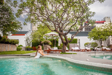 Young happy woman in white swimwear relax in swimming pool in modern tropical resort. Luxury vacation, Asia trip