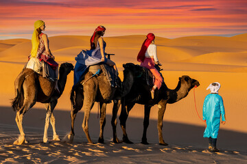 Three Riders And Their Handler Travel Through The Saharan Desert On Their Camels In Morocco