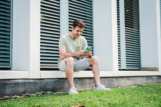 Young guy having fun playing a game with a cellphone sitting on the university campus after class. Teenage student smiling typing on a social media app with a smartphone device. High quality photo