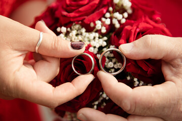 Braut und Bräutigam halten ihre Eheringe vor dem Brautstrauss mit roten Rosen - 559851995