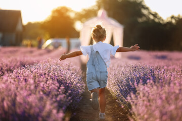 Back view of happy child girl runs raising her hands like flying plane in lavender field on summer warm day. Hyperactive little kid in sunglasses dreams of flying in nature. Children's fantasies.
