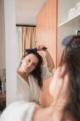 Fototapeta premium woman in a white bathrobe combing her hair with a brush in front of the mirror on her closet door in her bedroom